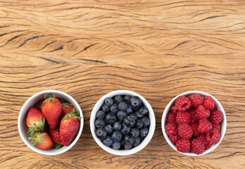 Raspberries, strawberries and blueberries in bowls over wooden table with copy space