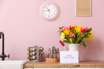 Jug with tulips, greeting card with text HAPPY WOMEN'S DAY and different utensils on kitchen counter near color wall