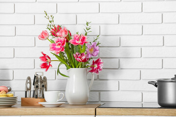 Jug with beautiful flowers and different utensils on kitchen counter near white brick wall