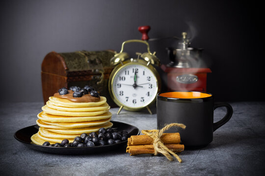 Pancakes with blueberries and chocolate paste on black dish, cup of coffee with steam, coffee grinder and cinnamon sticks, casket and alarm clock on gray table on black background