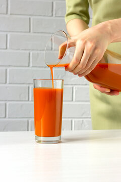 Woman holds a pitcher and is pours fresh carrot juice into glass in the kitchen. Healthy lifestyle and balance. Boost immunity. Delicious refreshment. Increase metabolism, help slimming. Vertical.