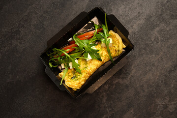 French omelette and tomato and arugula salad in a paper container on black background. Breakfast. Healthy food delivery concept.