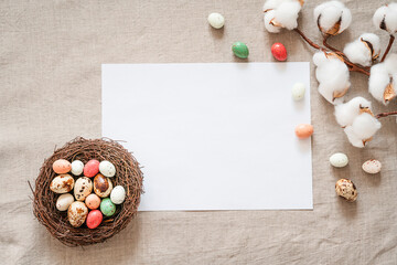 Easter background with white paper blank, cotton flowers, bird’s nest and colored easter eggs on a beige background. Easter composition on a textured beige linen background