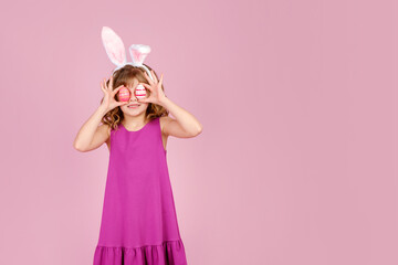Adorable smiling little girl with curly hair in fuchsia colored dress and bunny ear headband, smiling happily while covering eyes with eggs during Easter celebration, copy space