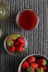 Overhead view of two uncovered glass jar. One full of strawberry marmalade, the other with some fresh strawberry pieces and lemon, next to a bowl full of more strawberries.