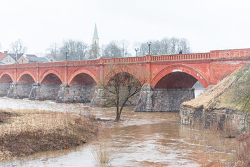 Fototapeta na wymiar Flood in spring day. Flooded road and tree next to old red brick bridge. Kuldiga, Latvia.