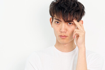 Portrait of handsome Chinese young man with curly black hair in white T-shirt posing against white wall background. Fingers on head and looking at camera, looks seriously, close up front view.