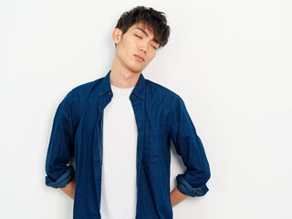 Portrait of handsome Chinese young man with curly black hair in blue shirt posing against white wall background. Hands on his back and eyes closed with tired expression, looks exhausted.