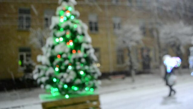 Christmas tree covered snow, glowing colorful lights during snowfall outdoors. Snowy city street with illumination decoration on winter night. Abstract Christmas New Year background. Light twinkling.