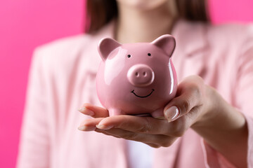 A cute girl in a pink jacket is holding a 3d piggy bank in her hands. Beautiful close-up portrait in studio on pink.