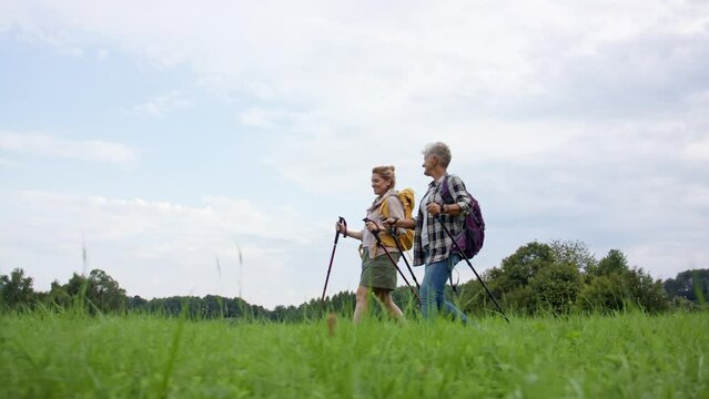 Low angle view of mid adult woman with trekking poles hiking with active senior mother outdoors in nature.