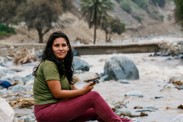 Woman sitting on a river bank holding a cell phone with her hands.