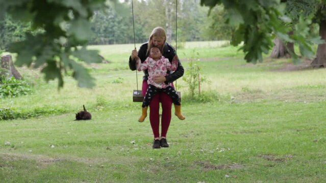 Biracial family tender moments, blonde European mother momy and little African girl daughter have fun playing happily on swing in garden, static shot through tree leaves, cloudy day