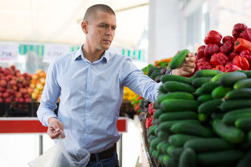 Caucasian man choosing cucumbers in greengrocer and putting them into plastic bag.