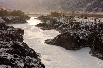 A narrow rocky channel of a frozen river flowing through a snow-covered steppe surrounded by mountains.