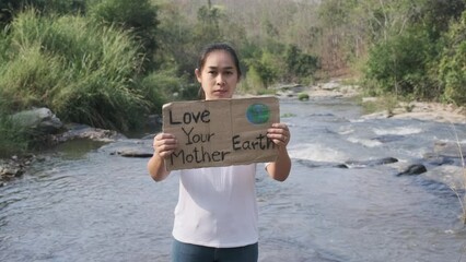 Portrait of woman standing with Love Your Mother Earth poster at a waterfall in the forest. Female volunteer activists demonstrate protecting the environment. World Environment Day