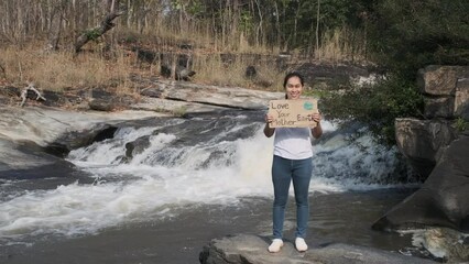 Portrait of woman standing with Love Your Mother Earth poster at a waterfall in the forest. Female volunteer activists demonstrate protecting the environment. World Environment Day