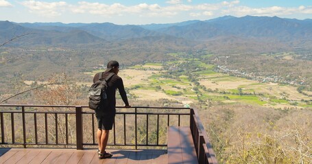 Fototapeta na wymiar Young tourist adventurous traveler Hiker man walking to balcony terrace and looking over tropical nature landscape in summertime sunny day in Thailand 