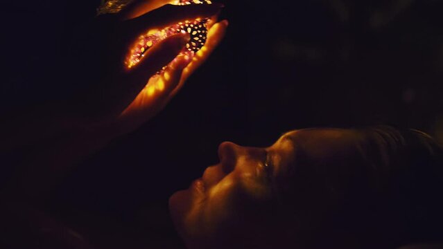 Magic hot light from lanterns falls on the woman eyes. The naked adult sexy caucasian girl lies at night on the bed and holds warm lamps in her hands. Christmas toy in black background. Indoor.