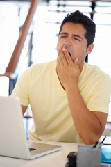 Time for my next cup of coffee. A young man yawning while sitting at a desk in front of a computer.