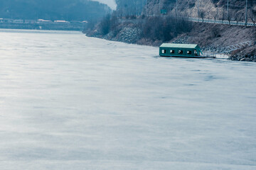 Green boathouse on frozen river in mountainous countryside.