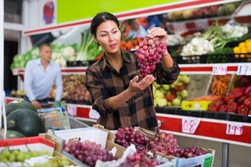Positive asian female buyer shopping in supermarket, choosing ripe purple grapes in fruit and vegetable department