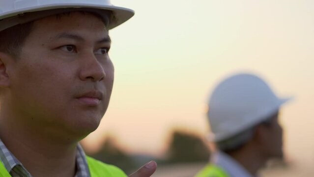 Close up, face of young engineer wearing white helmet while checking on operation of sun and cleanliness of photovoltaic solar panels at sunset