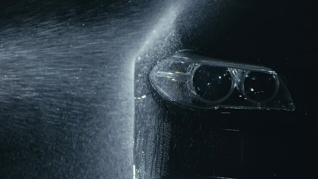 Car headlight wash. Washing modern vehicle body by high pressure jet wash hose water. Auto glass headlamp, angel eyes in drops. Close up of the spotlight of a black automobile as it gets cleaned.