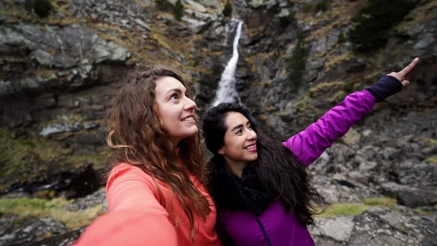 mixed race ethnicity best friend couple of woman sharing a video on social media taking selfie in natural mountains unpolluted landscape with rocky waterfall