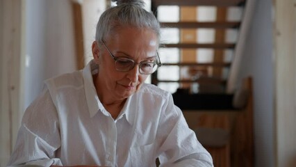 Thoughtful senior woman sitting at table and reading book at home.