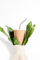 reusable eco cup and branches of plant in sleeve of sweater on white background. Sustainable lifestyle, conscious consumption, concept. copy space