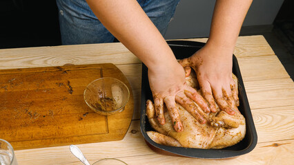 A close-up of a woman's hands smeared with seasoning and spices places a raw chicken in a black...