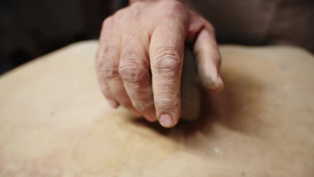 Mans wrinkled hand crumples and squeezes piece of clay on table, closeup. Craftsman processes piece of clay with his hands before working with it. Concept craft