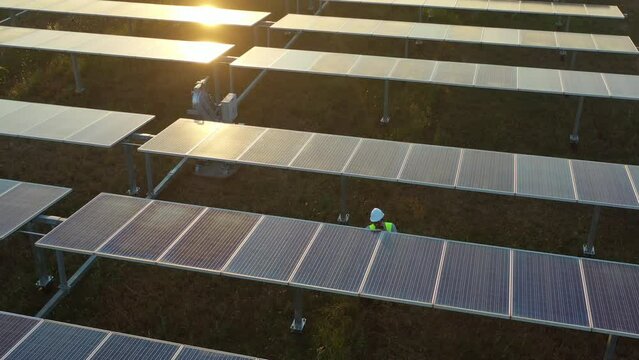 Aerial drone view, Flight over solar panel farm, young engineer wearing helmet checking on operation of sun and cleanliness of photovoltaic solar panels at sunset