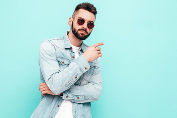 Portrait of handsome confident stylish hipster lambersexual model.Man dressed in jacket and jeans. Fashion male posing near blue wall in studio in sunglasses