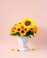 Collage with plaster antique sculpture of human face in a popart style. Creative concept ancient statue head, hair made of fresh sunflower flowers. Poster on pastel pink background.