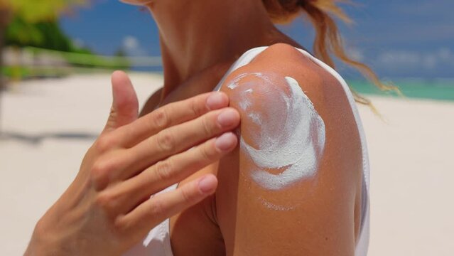 Close up of a happy smiling young woman is applying a sunscreen or sun tanning lotion on a shoulder to take care of her skin on a seaside beach during holidays vacation.