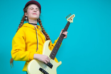 Dreaming Caucasian Teenager Guitar Musician Playing On Yellow Bass Guitar Posing In Fashionable Yellow Hoody Jacket Over Trendy Turquoise Background.