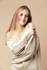 Happy Smiling Winsome Caucasian Blond Female Wearing Knitter Decorated Warm Sweater while Embracing Herself Over Beige Background