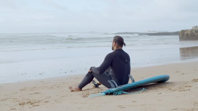 Tired surfer with artificial leg sitting on sandy beach. Back view of sportsman with bionic leg resting after surfing workout in ocean, looking at waves and relaxing. Sport, disability concept