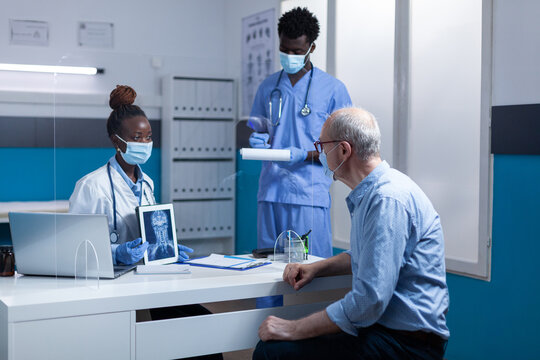 Clinic radiology specialist holding x-ray scan image and explaining illness to retired man while medical nurse taking notes on clipboard. Expert radiologist showing MRI scan image to elderly man.