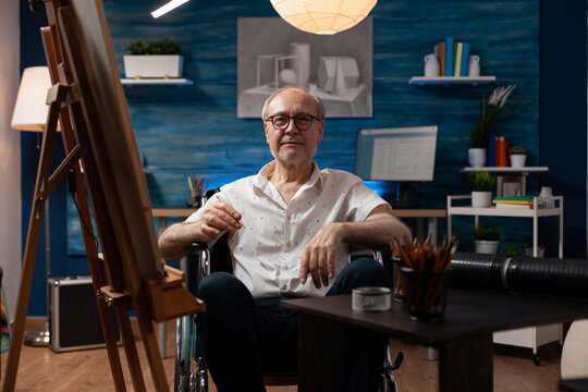 Elderly man sitting in wheelchair drawing black and white sketch using pencil on canvas in artist studio. Senior citizen who is paralyzed feeling creative in front of easel creating fine art.