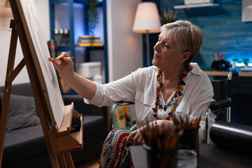 Portrait of elderly woman using wheelchair drawing static nature on easel in home art studio for...