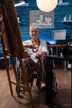 Portrait of senior painter holding pencil sitting in wheelchair in front of easel with canvas in drawing workshop. Confident retired sketch artist posing inspired in home hobby art studio.