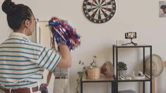 Rear-view shot of teenage cheerleader girl in striped polo t-shirt filming video on smartphone of herself dancing with pompons at home