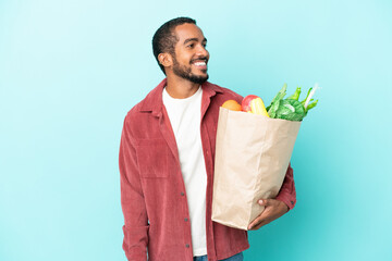 Young latin man holding a grocery shopping bag isolated on blue background looking side