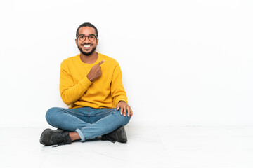 Obraz premium Young Ecuadorian man sitting on the floor isolated on white wall pointing to the side to present a product