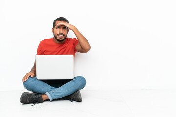 Young Ecuadorian man with a laptop sitting on the floor isolated on white background looking far away with hand to look something