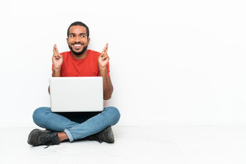 Young Ecuadorian man with a laptop sitting on the floor isolated on white background with fingers crossing