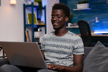 Freelancer man searching information for business lesson using laptop computer working remote from home. African american adult sitting on couch reading course for communication project